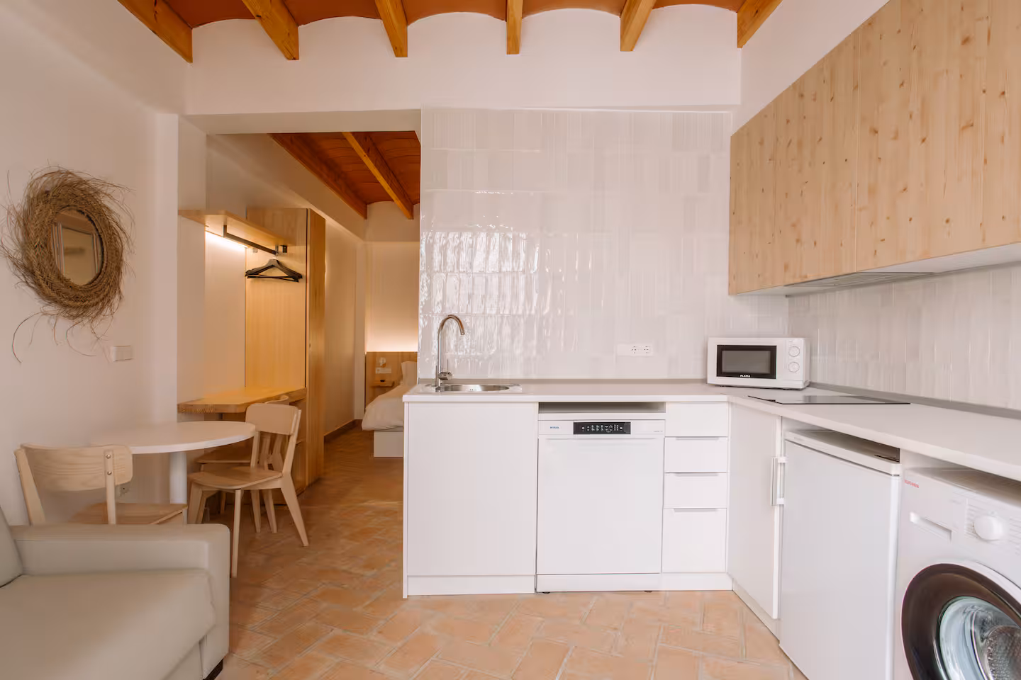 Cocina blanca bajo el techo abovedado de madera, con espejo circular de fibra y mesa de comedor