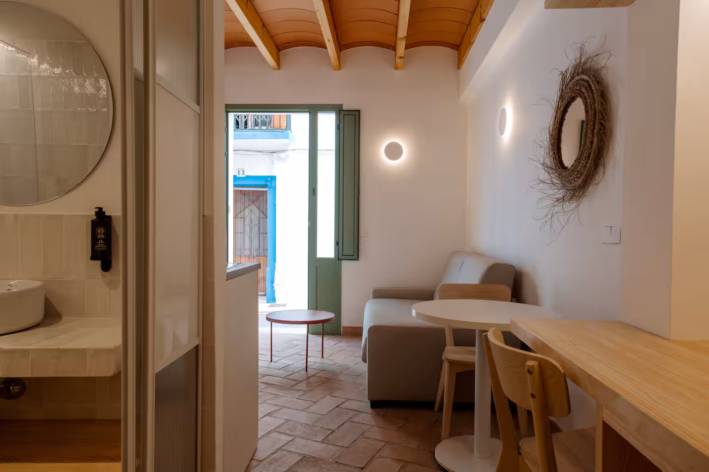 Salón de Termas Romanas con espejo circular de fibra natural, barra de madera y la puerta verde abierta a la calle